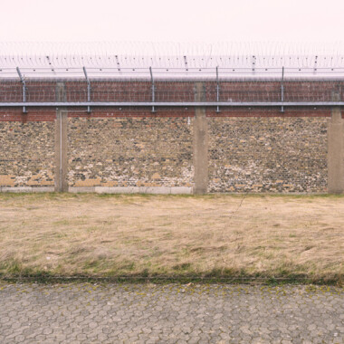 Die Außenmauer der JVA Rennelberg. Eine hohe Mauer aus Backstein und Stacheldraht umgibt eine freie Wiesenfläche.